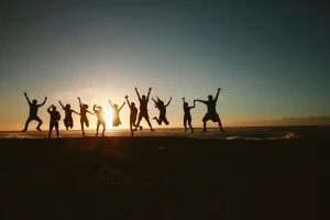 Grupo de personas saltando en una playa, esta atardeciendo y el sol da sombra a todas las personas
