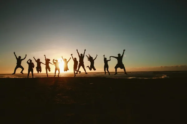 Grupo de personas saltando en una playa, esta atardeciendo y el sol da sombra a todas las personas