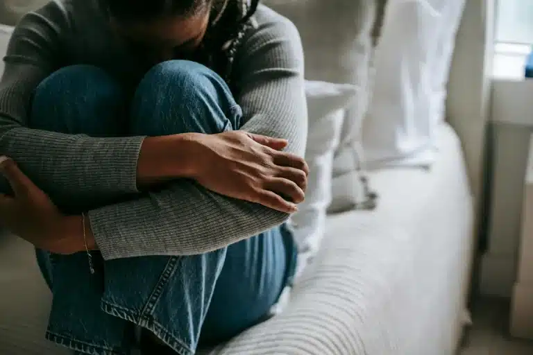 En la foto se observa a una mujer encima de la cama, abrazándose a las piernas y con la cabeza entre ellas.